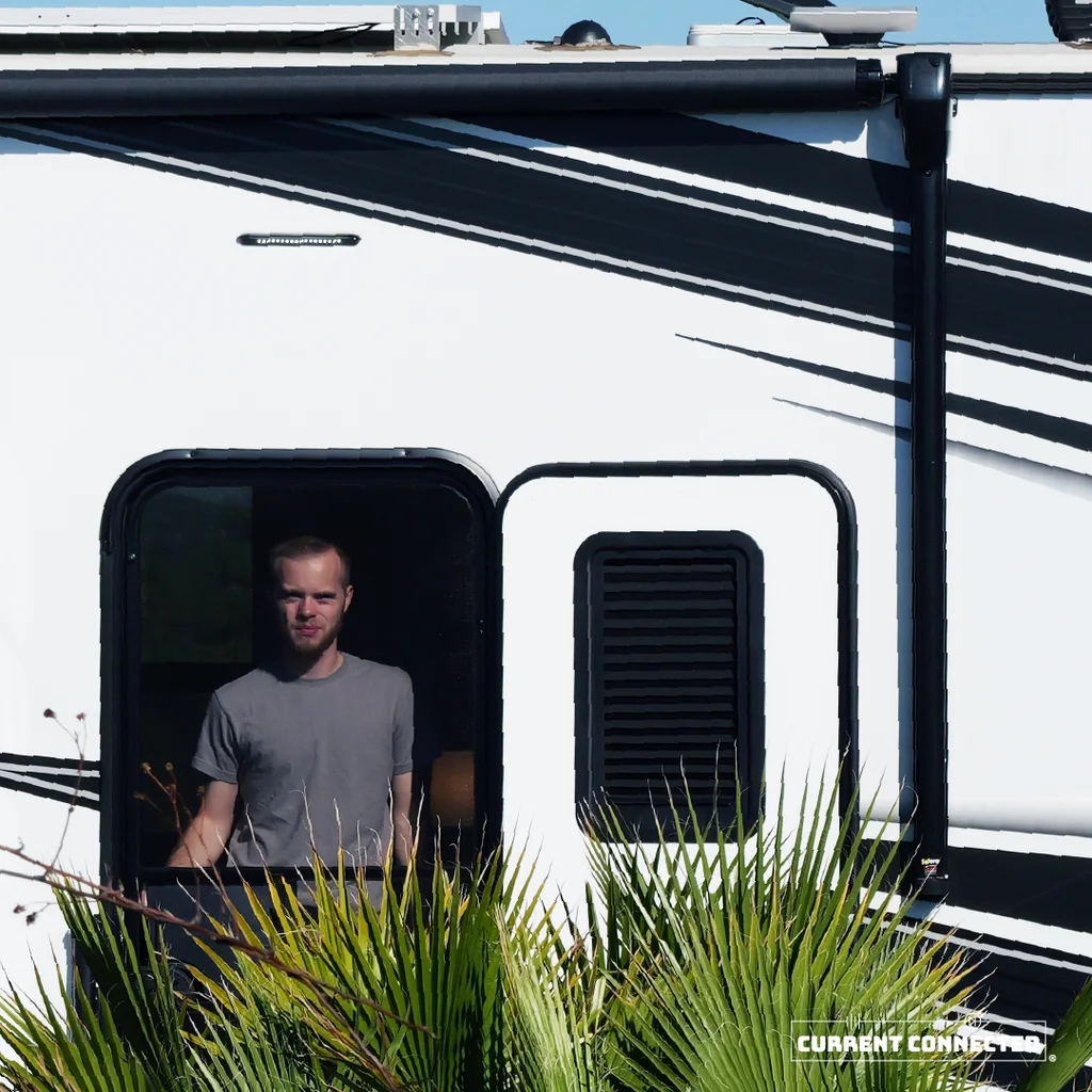 Photo of a man standing in the door of an RV. There are tropical plants in front of the RV door. 